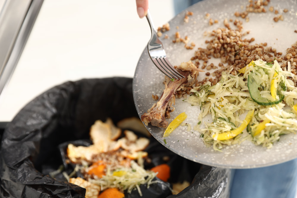 Food waste being scraped from a plate into a trash bin lined with a plastic bag.