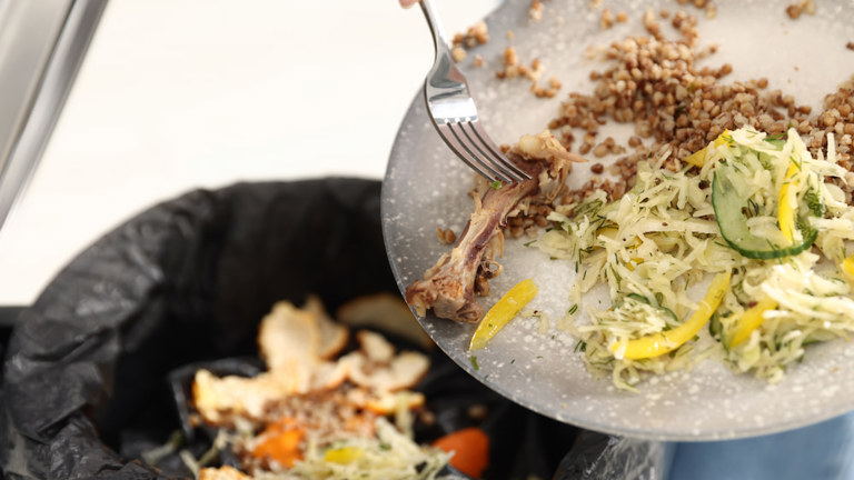 Food waste being scraped from a plate into a trash bin lined with a plastic bag.