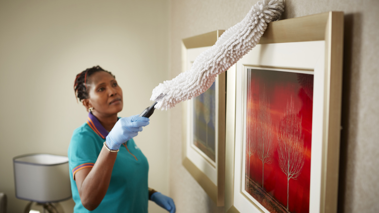 Housekeeper with red-brown braids in a teal uniform and blue gloves dusting a framed abstract picture with a gray duster.