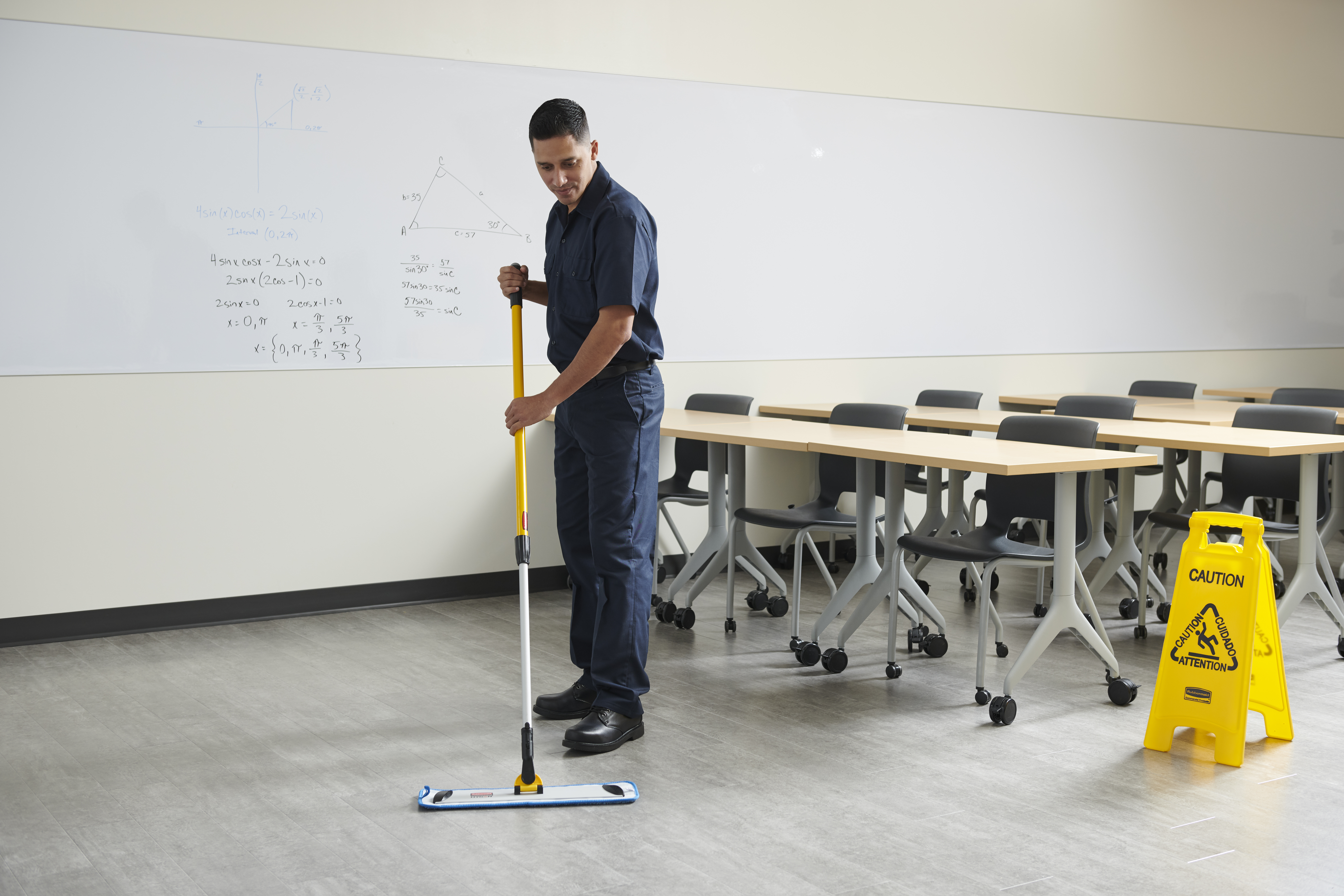 Male custodian cleaning the floor of an empty classroom with a flat mop, with furniture visible in the background.