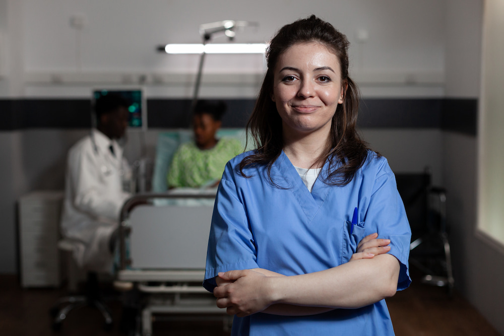 Confident nurse in blue scrubs standing with arms crossed in a hospital room, with a doctor and patient in the background.