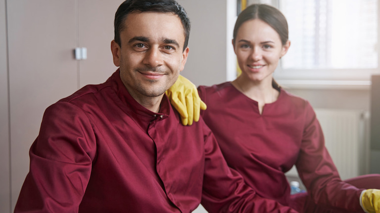 Smiling man and woman in matching dark red shirts and yellow gloves pose together, appearing to be professional cleaners or maintenance staff.